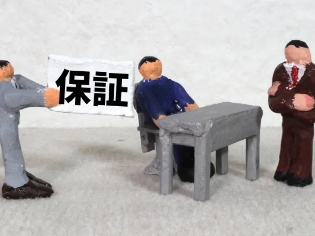 粘土でできたスーツ姿の人形が3体並んでおりそのうちの1体が保証と書かれた紙を持っている画像