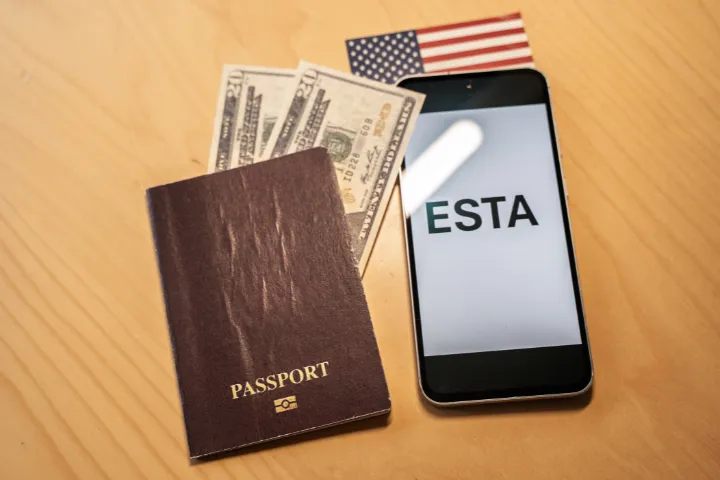 机の上にESTAの文字が表示されたスマホとアメリカ紙幣とアメリカ国旗とパスポートが置かれた画像