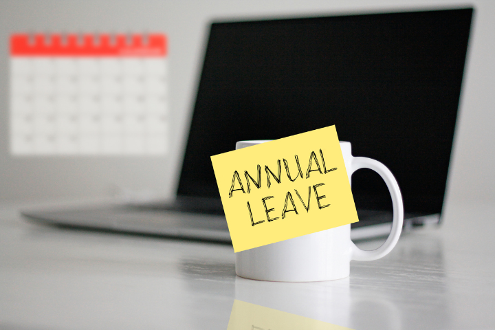 カレンダーとパソコンを背景にコップにANNUAL LEAVEと書かれた紙が貼られている画像